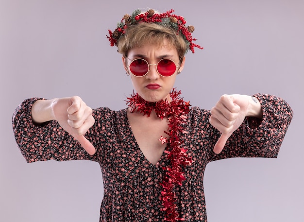 unpleased young pretty girl wearing christmas head wreath and tinsel garland around neck with glasses  showing thumbs down isolated on white wall