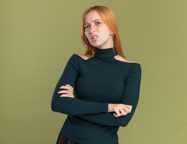 Unpleased young redhead ginger girl with freckles standing with crossed arms looking at camera on olive green