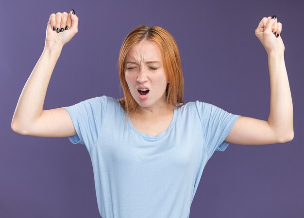 Unpleased young redhead ginger girl with freckles stands with closed eyes raising fists up