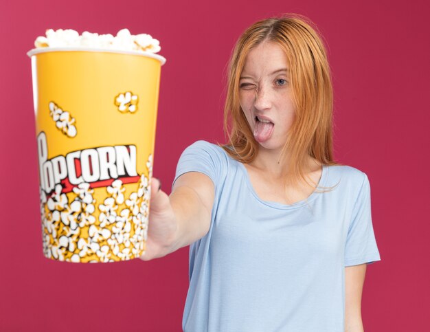 Una giovane ragazza rossa scontenta dello zenzero con le lentiggini sporge la lingua tenendo e guardando il secchio del popcorn isolato sulla parete rosa con lo spazio della copia