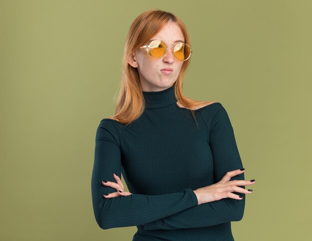 Unpleased young redhead ginger girl with freckles in sun glasses standing with crossed arms and looking at side on olive green