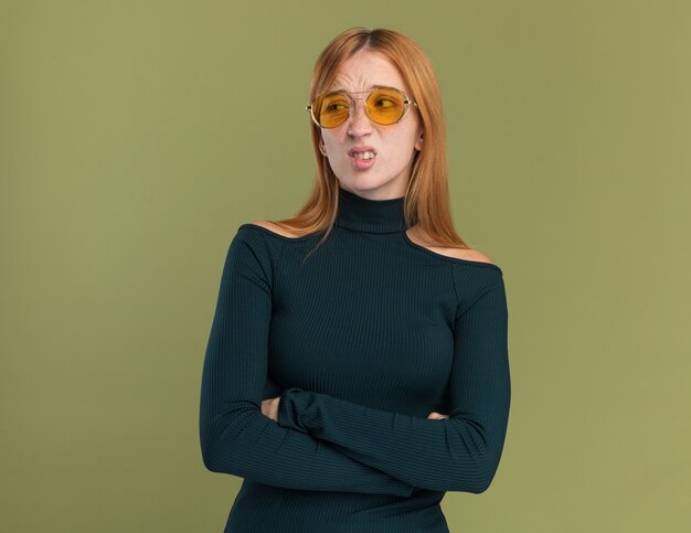 Unpleased young redhead ginger girl with freckles in sun glasses standing with crossed arms looking at side on olive green