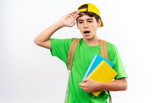 Unpleased young school boy wearing backpack with cap holding books putting hand on head 