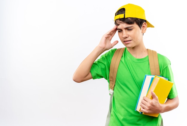 Unpleased young school boy wearing backpack with cap holding books putting hand on temple 