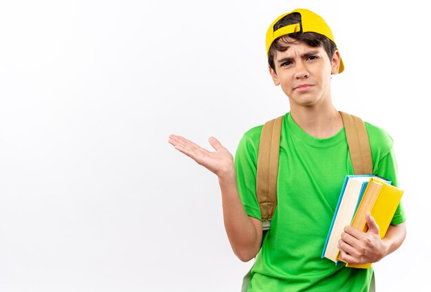 Unpleased young school boy wearing backpack with cap holding books spreading hand