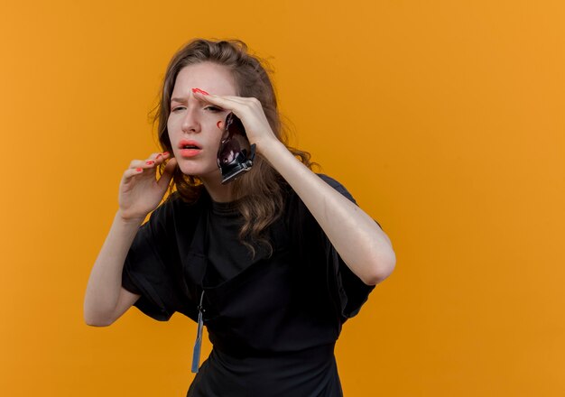 Unpleased young slavic female barber wearing uniform looking straight holding hair clippers keeping hands near face