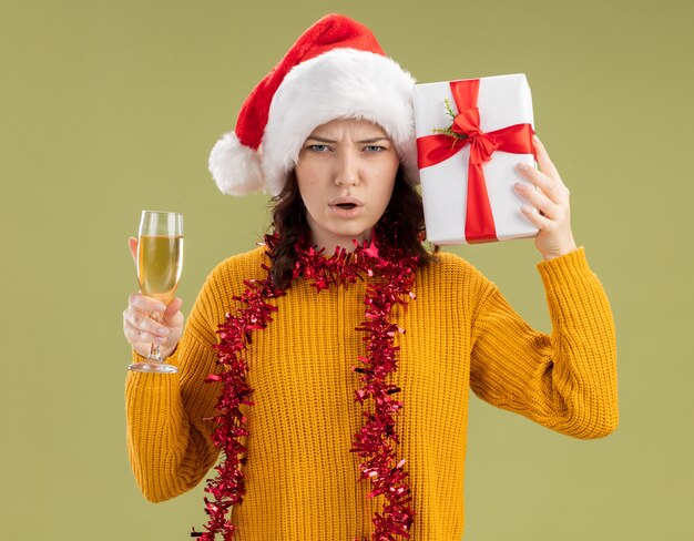 Недовольная молодая славянская девушка в шляпе санта-клауса и с гирляндой на шее держит бокал шампанского и рождественскую подарочную коробку, изолированную на оливково-зеленом фоне с копией пространства