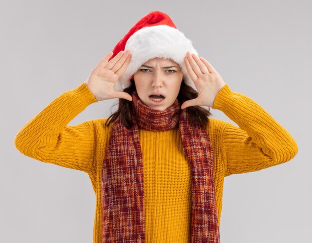 Unpleased young slavic girl with santa hat and with scarf around neck holds hands close to face isolated on white background with copy space