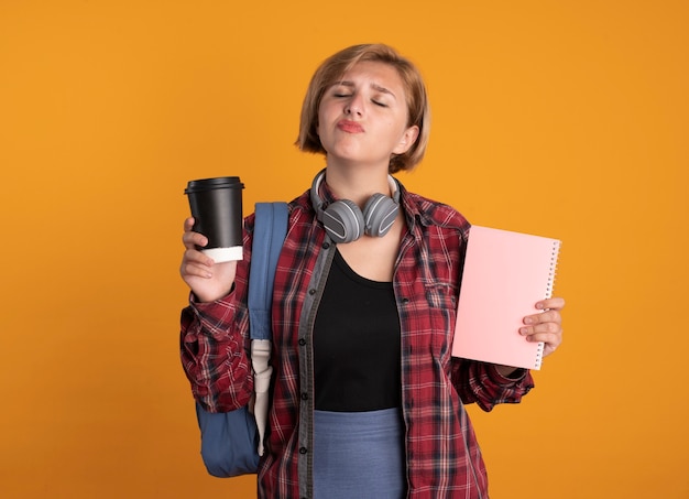 Unpleased young slavic student girl with headphones wearing backpack holds notebook and paper cup 