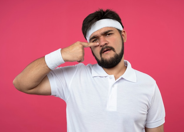 Недовольный молодой спортивный мужчина в повязке на голову и браслет, положив палец на нос, изолированный на розовом
