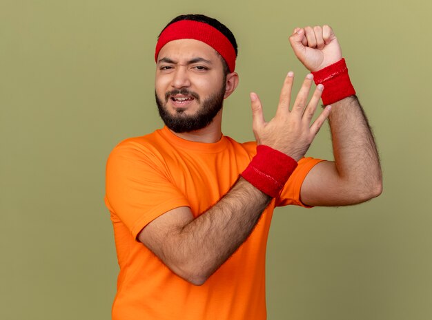 Unpleased young sporty man wearing headband and wristband raising fist with hand isolated on olive green background