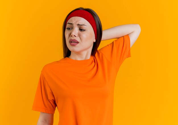 Unpleased young sporty woman wearing headband and wristbands looking at side keeping hand behind head 