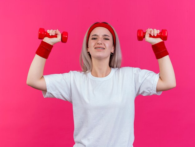 Недовольная молодая спортивная женщина с подтяжками, с повязкой на голову и браслетами стоит с поднятыми руками, держащими гантели, изолированными на розовой стене