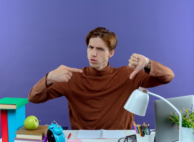 Unpleased young studend boy sitting at desk with school tools his thumb down and points at thumb on purple