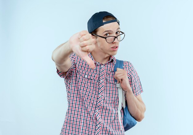 Unpleased young student boy wearing back bag and glasses and cap his thumb down on white