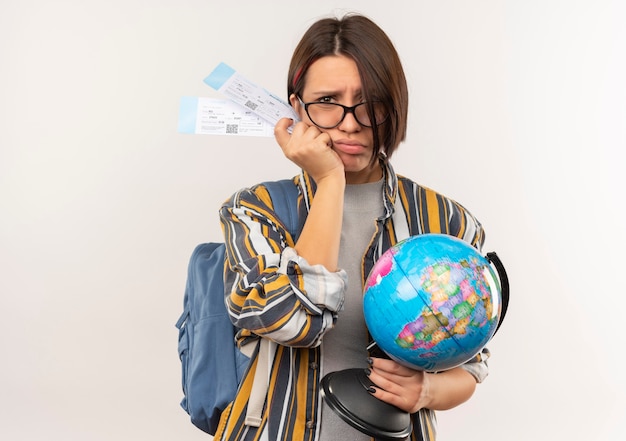 Unpleased young student girl wearing glasses and back bag holding airplane tickets and globe isolated on white background with copy space