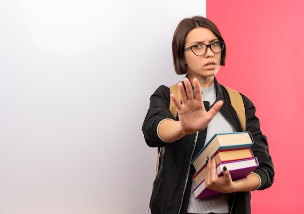 コピースペースでピンクの背景に孤立して身振りで示す白い壁の前に立っている本を保持している眼鏡とバックバッグを身に着けている不機嫌な若い学生の女の子