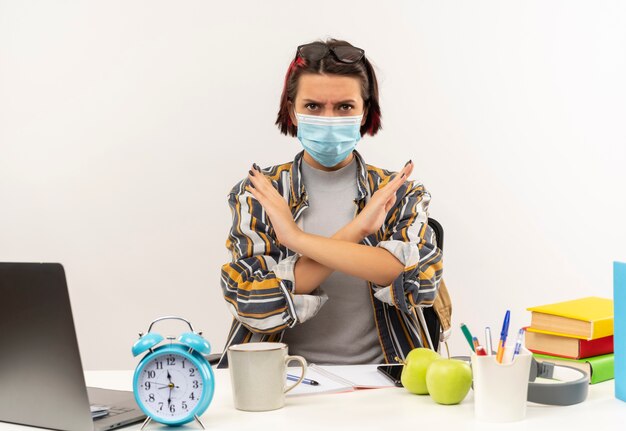 Unpleased young student girl wearing glasses on head and mask sitting at desk with university tools gesturing no isolated on white background