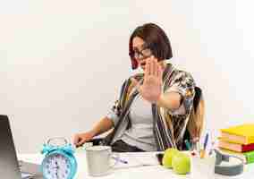 Free photo unpleased young student girl wearing glasses sitting at desk with university tools gesturing stop at camera isolated on white background