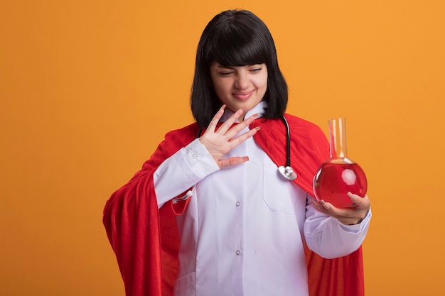 Unpleased young superhero girl wearing stethoscope with medical robe and cloak holding and looking at chemistry glass bottle filled with red liquid isolated on orange