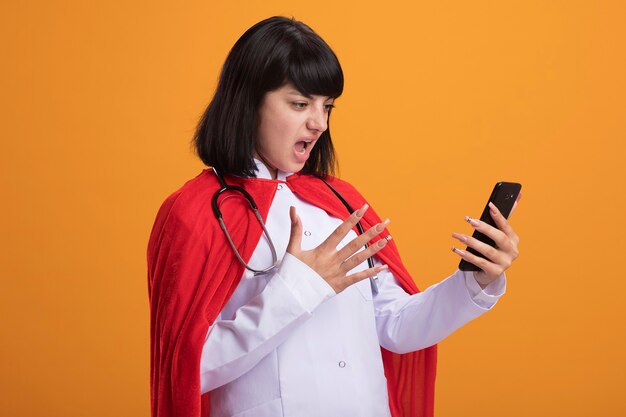 Недовольная молодая девушка-супергерой со стетоскопом в медицинском халате и плаще, держащая и смотрящая на телефон, изолированную на оранжевой стене