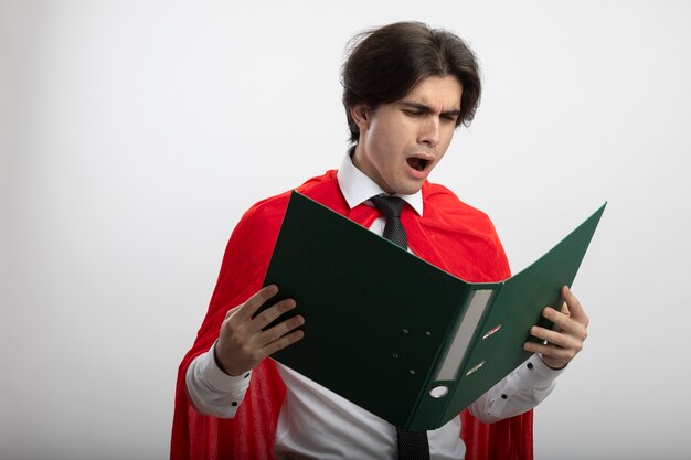 Unpleased young superhero guy wearing tie holding and looking at folder isolated on white