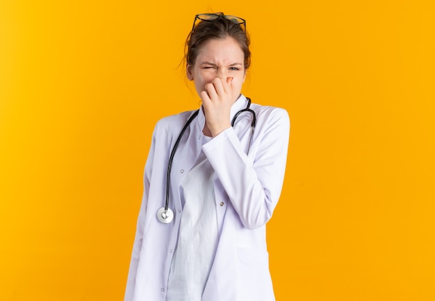 Unpleased young woman in doctor uniform with stethoscope closing her nose 