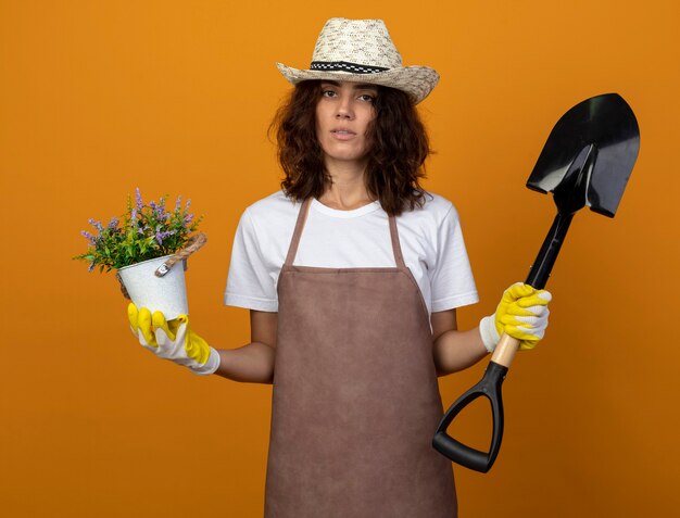 Unpleased young woman gardener in uniform wearing gardening hat and gloves holding flower in flowerpot and spade isolated on orange