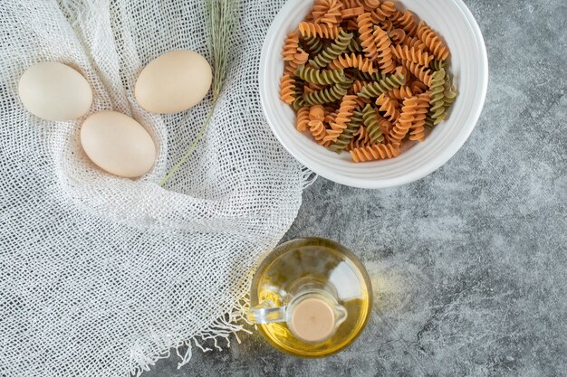 Unprepared spiral macaroni in white plate with three eggs and bottle of oil 