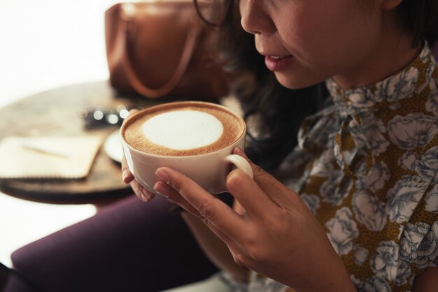 Unrecognizable Asian woman holding cup of cappuccino in cafe