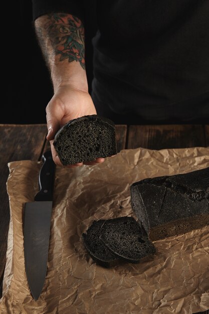 Unrecognizable baker with tattooed hands shows piece of freshly baked homemade charcoal bread, big chief knife near on craft paper on wooden rustic table ready for sale