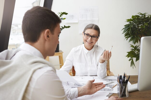 Unrecognizable brunette young male architect sitting at desk with drawings while discussing something with his cheerful mature female boss who is smiling at him, approving his creative ideas