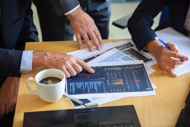 Free photo unrecognizable businessman pointing at printed chart and showing graph to colleagues. professional content partners making notes for statistics. cooperation, communication and partnership concept