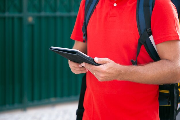 Unrecognizable courier standing and holding tablet in hands. Cropped view of deliveryman delivering order in thermal backpack and wearing red shirt. Delivery service and online shopping concept