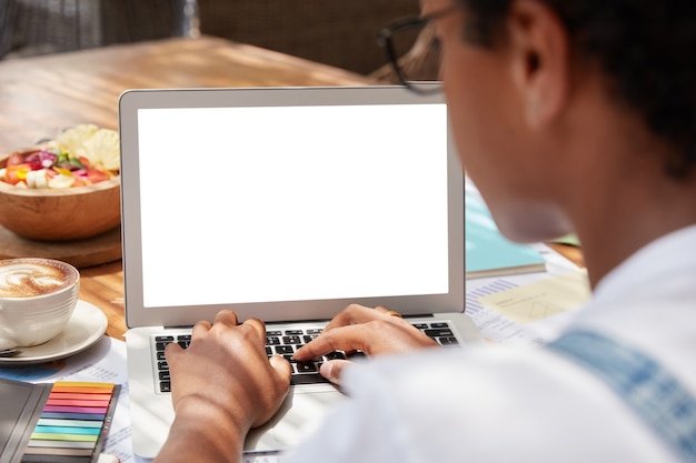 Unrecognizable dark skinned woman keyboards on laptop computer with blank screen area for advertising content