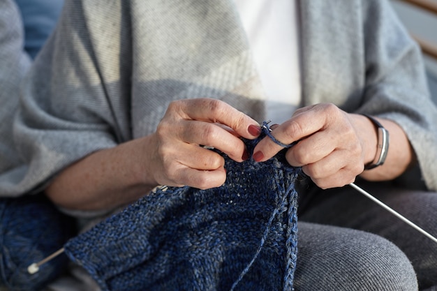 Unrecognizable elderly senior woman wearing wide gray scarf and wrist watch, knitting sweater. Close up view of aged female hands holding needles and yarn, doing needlework. Selective focus