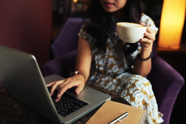 Неузнаваемая элегантная женщина, держащая капучино и работающая на ноутбуке в кафе