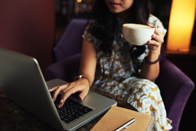 Unrecognizable elegant woman holding cappuccino and working on laptop in cafe