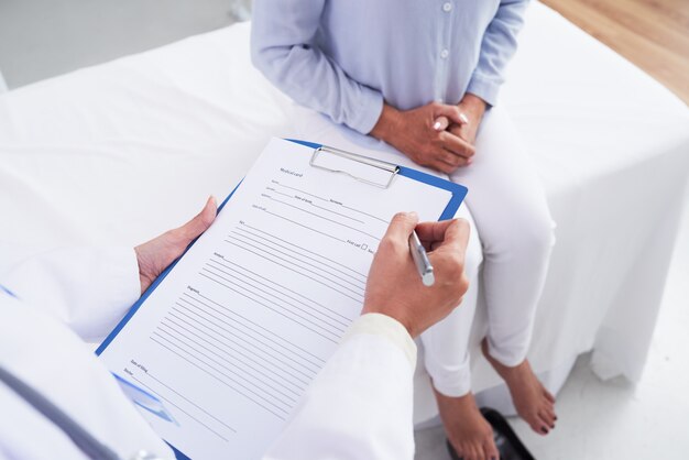 Unrecognizable female doctor filling in document and patient sitting on couch