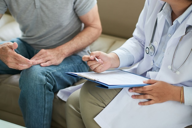 Unrecognizable female doctor sitting on couch with male patient and filling form