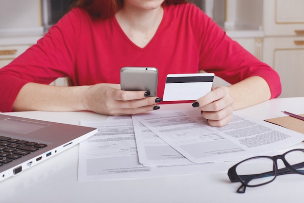 Foto gratuita il modello femminile irriconoscibile in maglione rosso si siede al tavolo di lavoro circondato da documenti e computer portatile