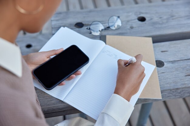 Unrecognizable female writer puts down new chapter of book in notepad notes some records holds mobile phone with blank screen poses on wooden bench creats organisation plan uses pen