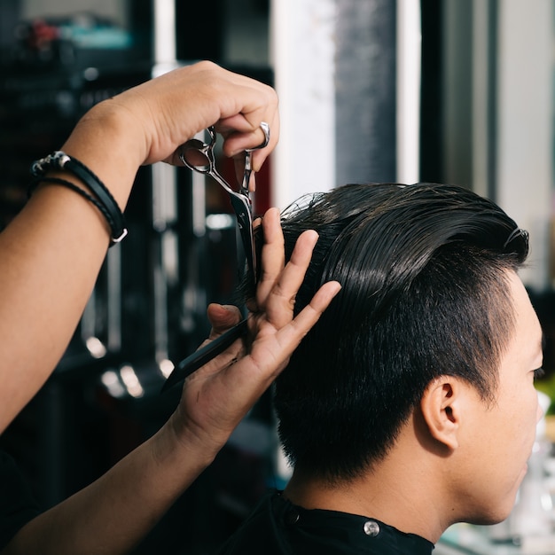 Unrecognizable hairdresser cutting Asian male customer's hair with scissors in salon