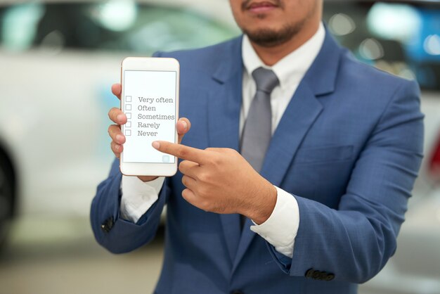 Unrecognizable man in business suit holding up smartphone and pointing to survey on screen