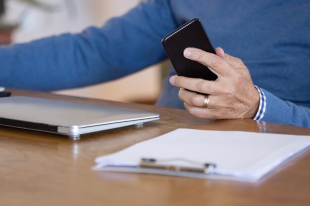 Unrecognizable man holding phone while sitting at table at home. Close-up shot of mature man working in home office, using smartphone and laptop, talking with colleagues. Digital device, workplace con