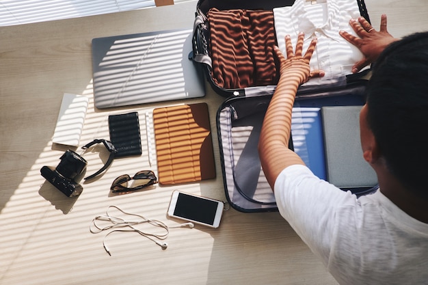 Unrecognizable man packing suitcase for trip, and electronic gadgets lying nearby