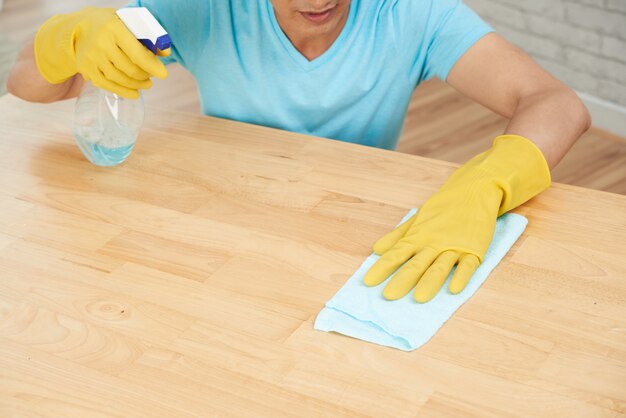 Unrecognizable man in rubber gloves spraying table and cleaning with cloth