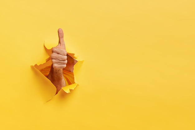 Unrecognizable man shows like gesture through torn yellow wall, keeps thumb up