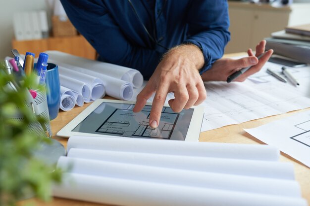 Unrecognizable man sitting at desk with technical drawings and looking at floor plan on tablet