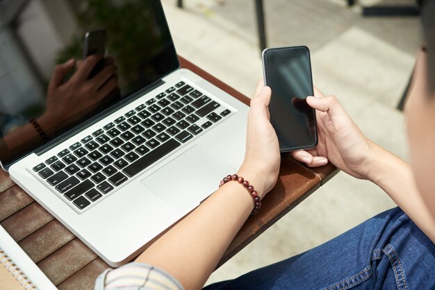 Unrecognizable man sitting with laptop in cafe and using smartphone